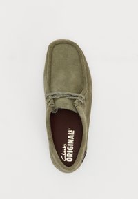 Chaussure en daim vert olive à bout rond, avec des coutures décoratives et des lacets ornementaux. L'intérieur présente une semelle brune avec le texte "Clarks Originals".