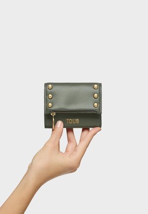 Ręka trzymająca mały oliwkowozielony skórzany portfel z złotymi ćwiekami, zamkiem błyskawicznym i logo "TOUS" na jednolitym jasnoszarym tle.