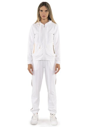 Une femme porte un sweat à capuche blanc zippé et un pantalon de jogging assorti aux détails dorés, ainsi que des baskets blanches, sur un fond uni.