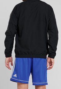 Svart idrottsjacka med lång ärm, elastisk nederkant och vita sidosträck, parat med blå shorts som har vit piping och en Adidas-logotyp.