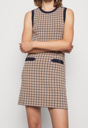 Robe sans manches beige et bleu marine à carreaux avec bordure bleu marine sur le col et les poches, portée par une personne les bras croisés.
