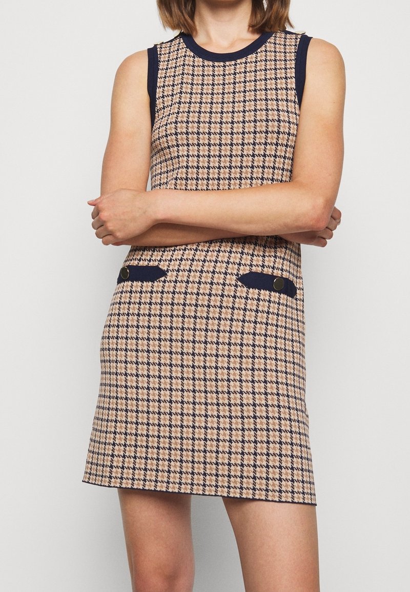 Robe sans manches beige et bleu marine à carreaux avec bordure bleu marine sur le col et les poches, portée par une personne les bras croisés.