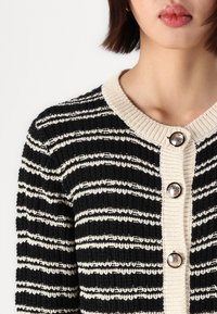 Zwart en crèmekleurig gestreepte gebreide cardigan met een geribde textuur, voorzien van een ronde halslijn en drie decoratieve knopen aan de voorkant.