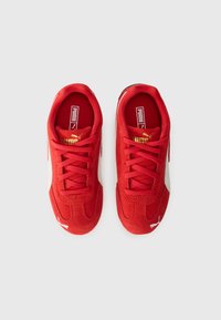 Baskets de sport en suède rouge avec des accents en cuir blanc, forme arrondie, surface texturée et détail en or sur la langue et le côté.