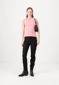 Rózsaszín, testhez simuló póló fekete logóval, fekete, slim-fit farmernadrággal és fekete magassarkú csizmával párosítva. A modellen egy kisméretű fekete kézitáska van.