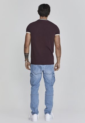 Mężczyzna stojący tyłem, ubrany w ciemnoczerwony t-shirt, jasnoniebieskie jeansy typu cargo i białe trampki, z tatuażami i bransoletkami na ramionach.