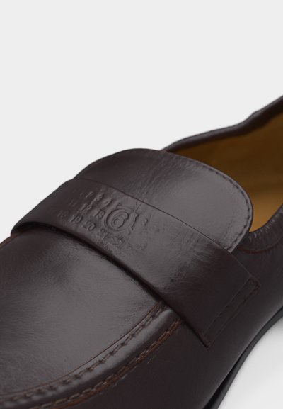 Σκούρο καφέ δερμάτινο μοκασίνι με ανάγλυφη διακοσμητική λωρίδα και εμφανές ράψιμο κατά μήκος της περιοχής των δακτύλων.