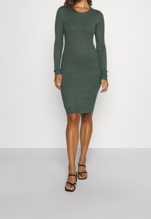 Femme portant une robe ajustée à manches longues côtelée, de couleur vert foncé, arrivant aux genoux, et des sandales noires à lanières, debout sur un sol gris.