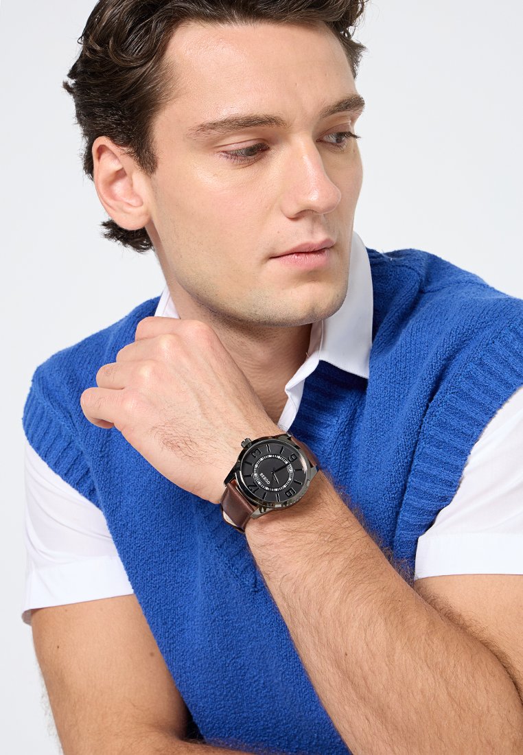 Reloj redondo negro con correa de cuero marrón, con esfera texturizada y detalles plateados. Llevado en la muñeca de una persona con un chaleco de punto azul.