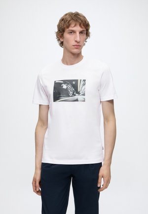 Νέος άνδρας που φοράει λευκό t-shirt με ασπρόμαυρο τύπωμα ενός τροχού αυτοκινήτου σε κίνηση, συνδυασμένο με σκούρο παντελόνι.
