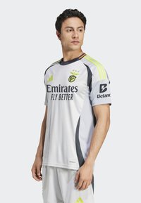 Sportstrøje i lys grå med sorte accenter, prydet med "Emirates" logo, tre striber på skuldrene og "Betano" logo på ærmet.