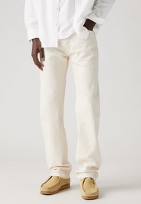 Hombre vistiendo pantalones rectos de color blanco roto, zapatos mocasín de ante color tostado, y una camisa blanca de botones con una mano en el bolsillo.
