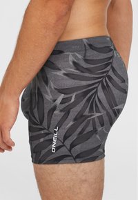 Čierne plavky s šedým vzorom listov, vyrobené z elastického materiálu, s bielym logom na boku. Priliehavý strih s hladkou textúrou.