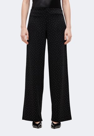 Mannequin portant un pantalon noir à jambes larges, orné de petits pois blancs, et des chaussures noires à bout pointu.