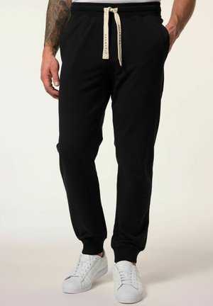 Pantalones de chándal negros de tejido suave, con cinturilla elástica y cordón, y bolsillos laterales. Combinados con zapatillas blancas.