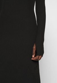 Gros plan d'une main dans une robe en maille côtelée noire à manches longues avec une ouverture pour le pouce au niveau du poignet sur un fond uni.
