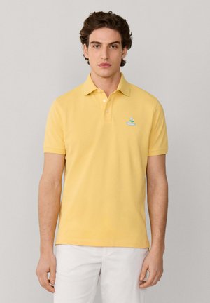 Joven vestido con una camiseta polo amarilla de manga corta con un pequeño logo azul claro en el pecho y pantalones blancos, de pie frente a un fondo gris.