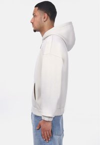 Un sweat à capuche gris clair avec une poche kangourou, des poignets côtelés et une coupe décontractée, porté avec un jean en denim bleu clair, présenté de profil.