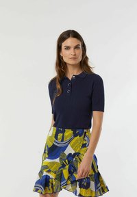 Navy geribbelde gebreide polo top met korte mouwen en drie knopen, gecombineerd met een veelkleurige uitlopende rok met gele en blauwe bloemenpatronen.