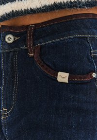 Tmavě modré džíny z denimu s hnědým detailem na kapse, zlatým prošíváním, kovovým knoflíkovým zapínáním a štítkem s grafikou řasy.