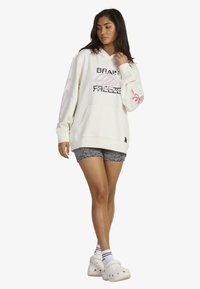 Cream-colored hoodie with black and pink graphic text. Features a front pocket, drawstring hood, and ribbed cuffs. Paired with striped shorts.