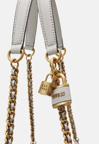 Valkoinen nahkainen olkalaukkukahva, jossa on kultaiset metalliketjut ja koristeet. Sisältää lukkon, jossa on kaiverrettuna "GUESS" ja "EST 1981".