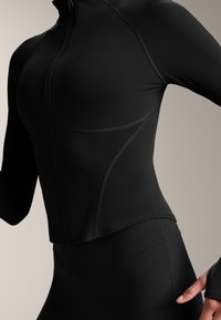 Černá sportovní bunda s dlouhými rukávy a zipem, která má prošívané detaily a přiléhavý střih. Vyrobena z hladkého, elastického materiálu.