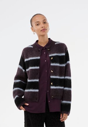 Strikket cardigan med en fluffy tekstur, mørk lilla og sorte striber med lyseblå accenter samt gyldne knapper. Laget over en mørk skjorte.