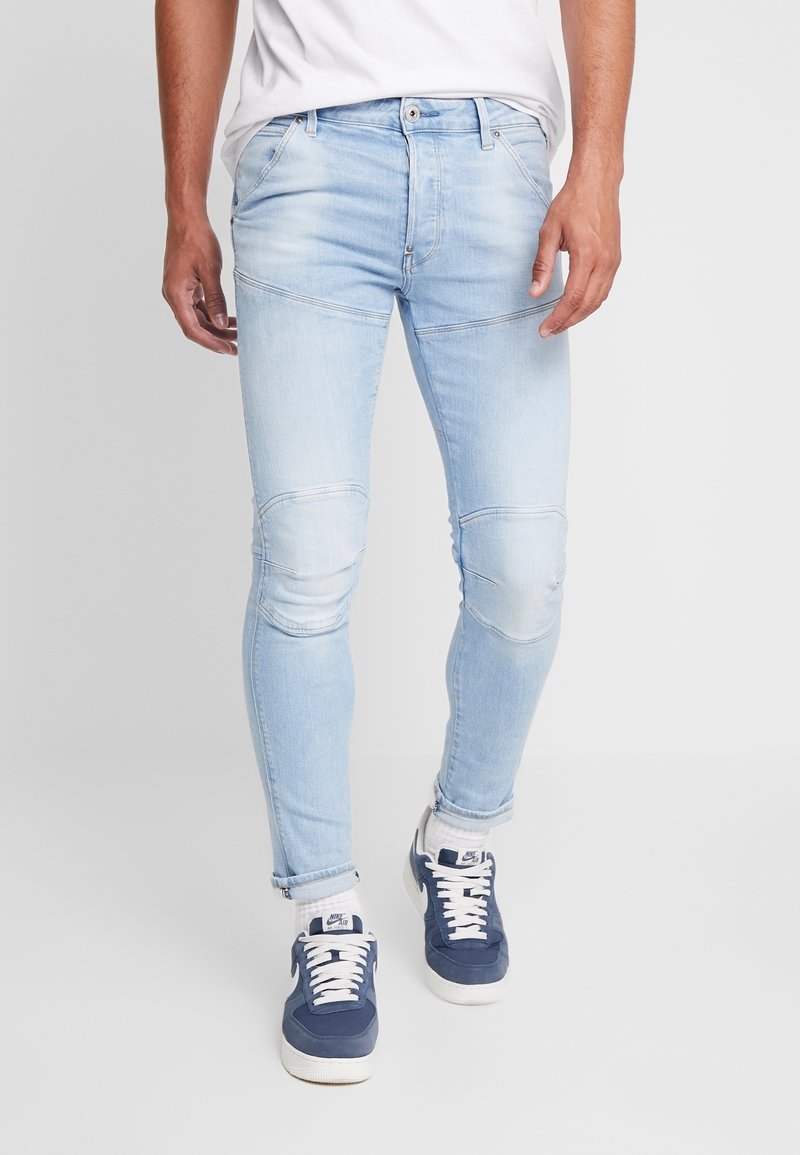 Homme portant un jean slim bleu clair avec des détails de couture au genou, des chaussettes blanches, des baskets Nike Air bleu marine et blanches, et un t-shirt blanc.