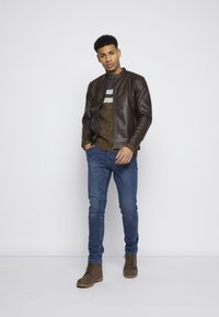 Chaqueta bomber de cuero marrón sobre un suéter estampado, jeans azules y botas marrones con cordones. El modelo está de pie contra un fondo blanco liso.