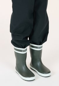 Bottes de pluie en caoutchouc vert avec des rayures blanches en haut, semelles texturées et un logo de marque sur le revers. Portées avec un pantalon imperméable noir.