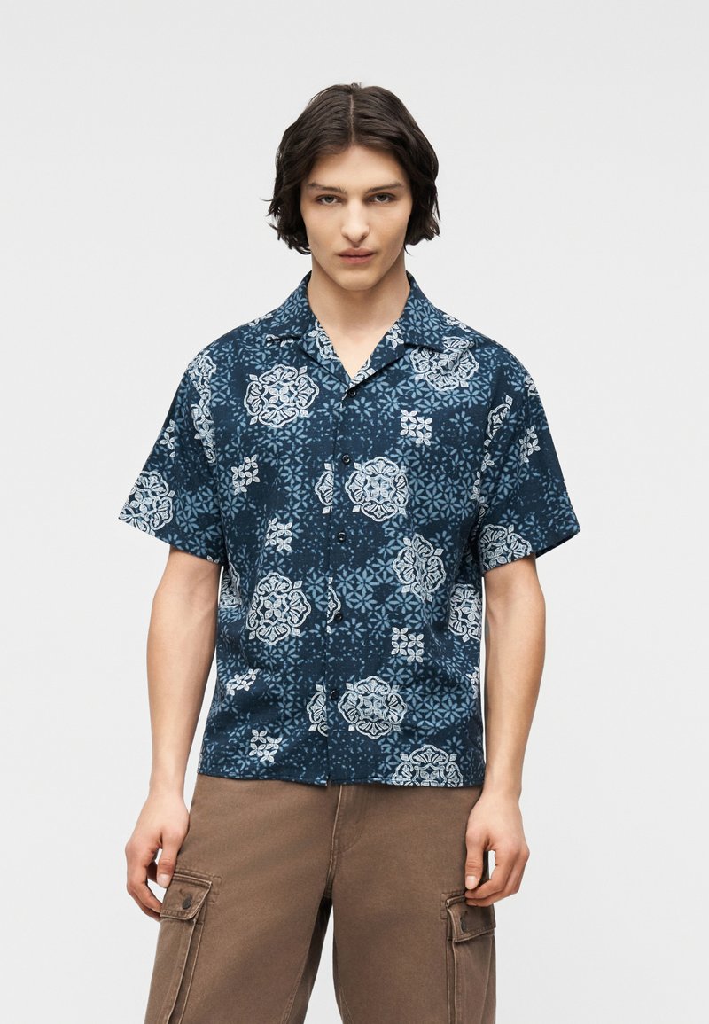 Νεαρός άνδρας με μεσαίου μήκους σκούρα μαλλιά, που φοράει μπλε πουκάμισο με κοντά μανίκια και σχέδιο, καθώς και καφέ παντελόνι cargo, σε λευκό φόντο.