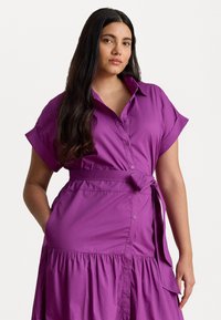 Violetti mekko, jossa on lyhyet käännettävät hihat, nappikiinnitys edessä ja vyötärönauha. Mekossa on kerroksellinen hameosa ja sivutaskut.
