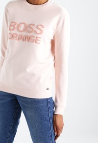 Suéter de color rosa claro hecho de una tela suave, con el texto elevado "BOSS ORANGE" en un color a juego. Escote clásico tipo crew y puños acanalados.
