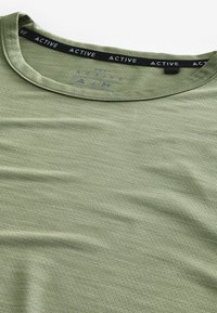 Grøn aktiv t-shirt med rund halsudskæring, struktureret stof og et kontrastlogo-bånd på kraven. Glat finish, letvægtsdesign.