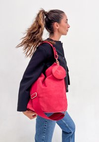 Femme avec une longue queue de cheval portant une veste noire et un jean, portant un grand sac à bandoulière en daim rouge avec une pochette ronde sur fond uni.
