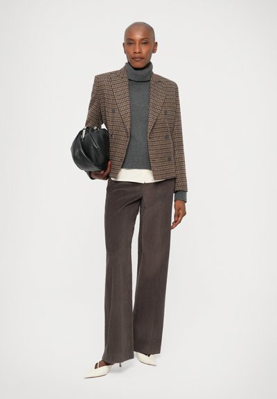 Brązowy blazer w kratkę houndstooth, szary sweter z golfem, biała koszula, brązowe szerokie spodnie z korduroyu oraz białe czółenka w szpic. Czarna torba w ręku.