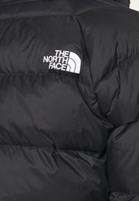 Doudoune noire avec le logo blanc "The North Face" sur la poitrine, montrant des coutures détaillées et une texture matelassée.