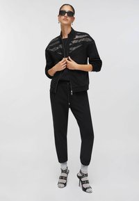 Veste noire à fermeture éclair avec un motif à pois sur les épaules, associée à un pantalon noir. Sandales à talons hauts avec des brides et des chaussettes à motifs complètent la tenue.