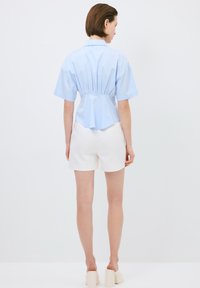 Camicia azzurre chiara a maniche corte con vita arricciata, orlo leggermente curvo, indossata con pantaloni corti bianchi. Texture liscia e design pulito.