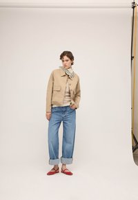 Jeune femme debout portant une veste beige, un chemisier à motifs, un jean bleu retroussé, des ballerines rouges et un foulard noir et blanc à pois autour du cou, en studio.