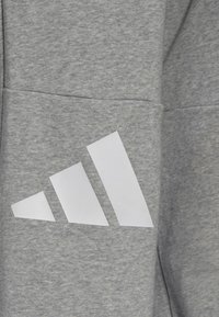 Grauer Sweatshirt-Stoff mit glatter Struktur, versehen mit einem auffälligen weißen Drei-Streifen-Logo-Design im Bereich der linken Tasche.