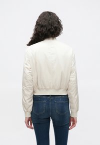 Blouson aviateur crème à coupe courte, avec poignets côtelés et tissu lisse, associé à un jean bleu. Le fond est blanc uni.