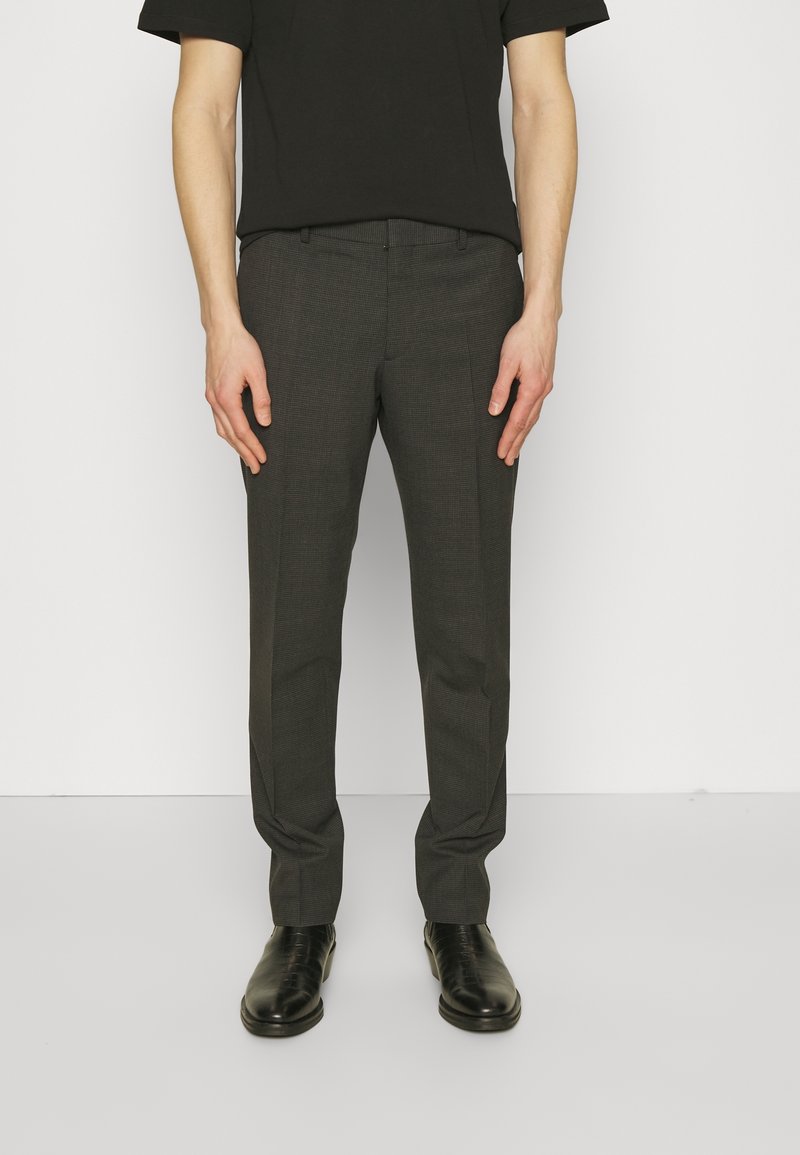 Homme portant un pantalon de costume gris foncé, une chemise noire à manches courtes et des chaussures en cuir noires, debout devant un fond blanc.