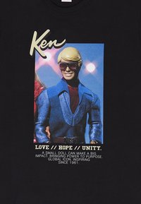 Schwarzes T-Shirt mit einem Grafikdesign von einer Ken-Puppe in einem blauen Anzug mit großen Sonnenbrillen, begleitet von Texten, die Liebe, Hoffnung und Einheit betonen.