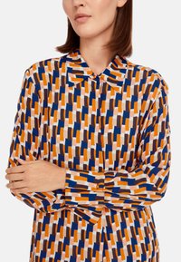 Mehrfarbige Bluse mit einem geometrischen Muster in Orange, Marineblau, Braun und Weiß, mit spitzen Kragen und knöpfbaren Manschetten.