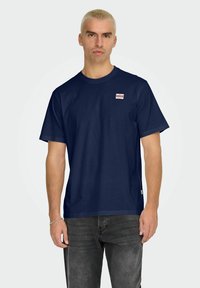 Marineblå bomulds t-shirt med korte ærmer, rund halsudskæring og et lille rødt og hvidt logo på venstre bryst. Struktureret stof.