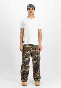 Tricou alb cu mânecă scurtă purtat împreună cu pantaloni cargo în tonuri de verde și maro cu model de camuflaj, și cizme crem. Șapcă cu un model de camuflaj.