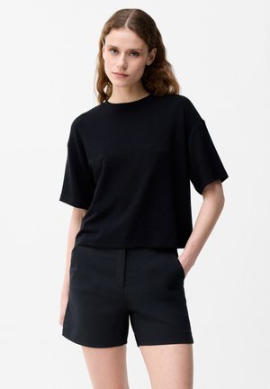 Młoda kobieta z falowanymi włosami, ubrana w luźny czarny krótki rękaw i czarne, dopasowane szorty, stoi z jedną ręką w kieszeni.