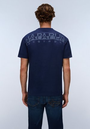 Mand med krøllet hår set bagfra iført en mørkeblå t-shirt med falmet tekst og mørkeblå jeans med påsyede lommer.